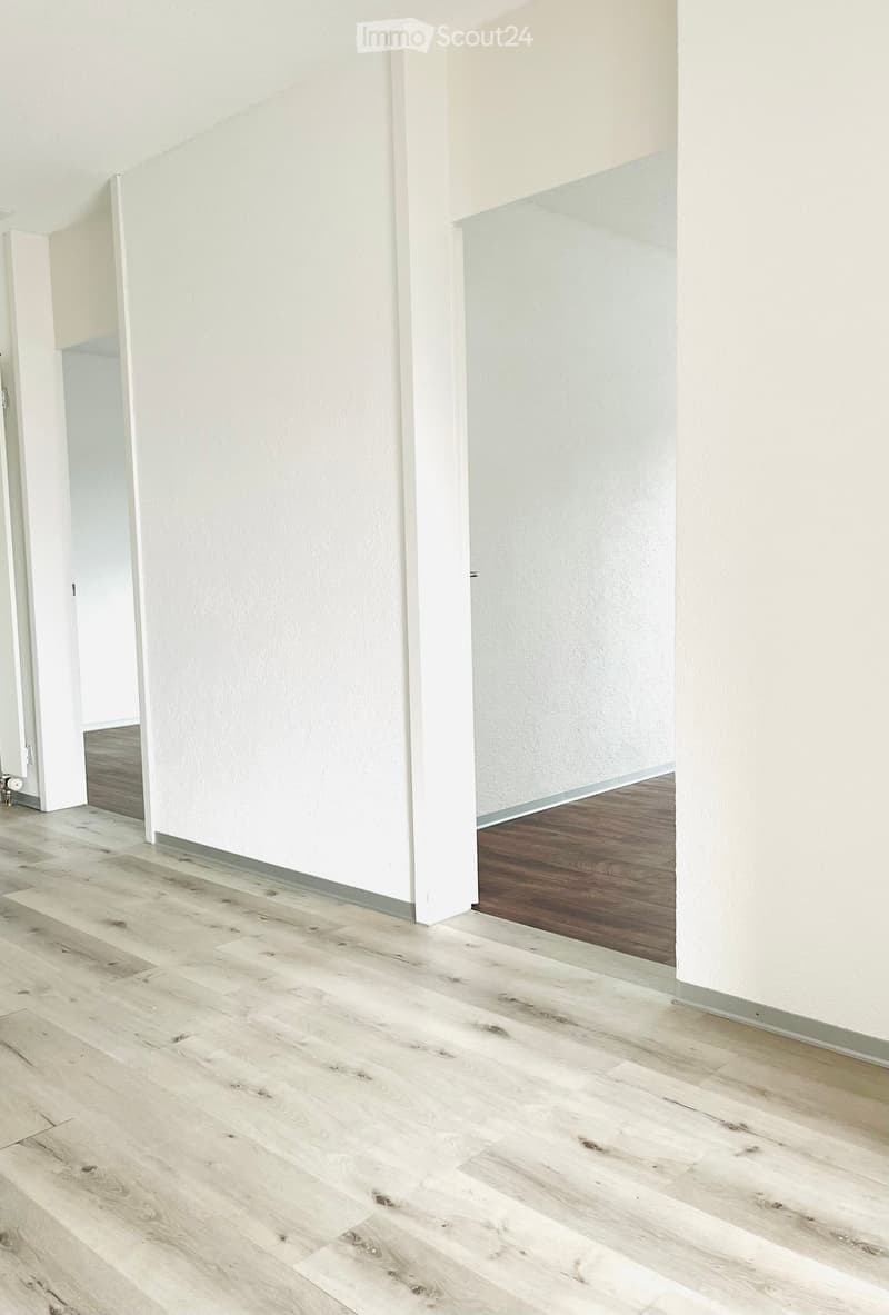 Empty room with light wooden flooring and white walls, featuring an open doorway leading to another room.