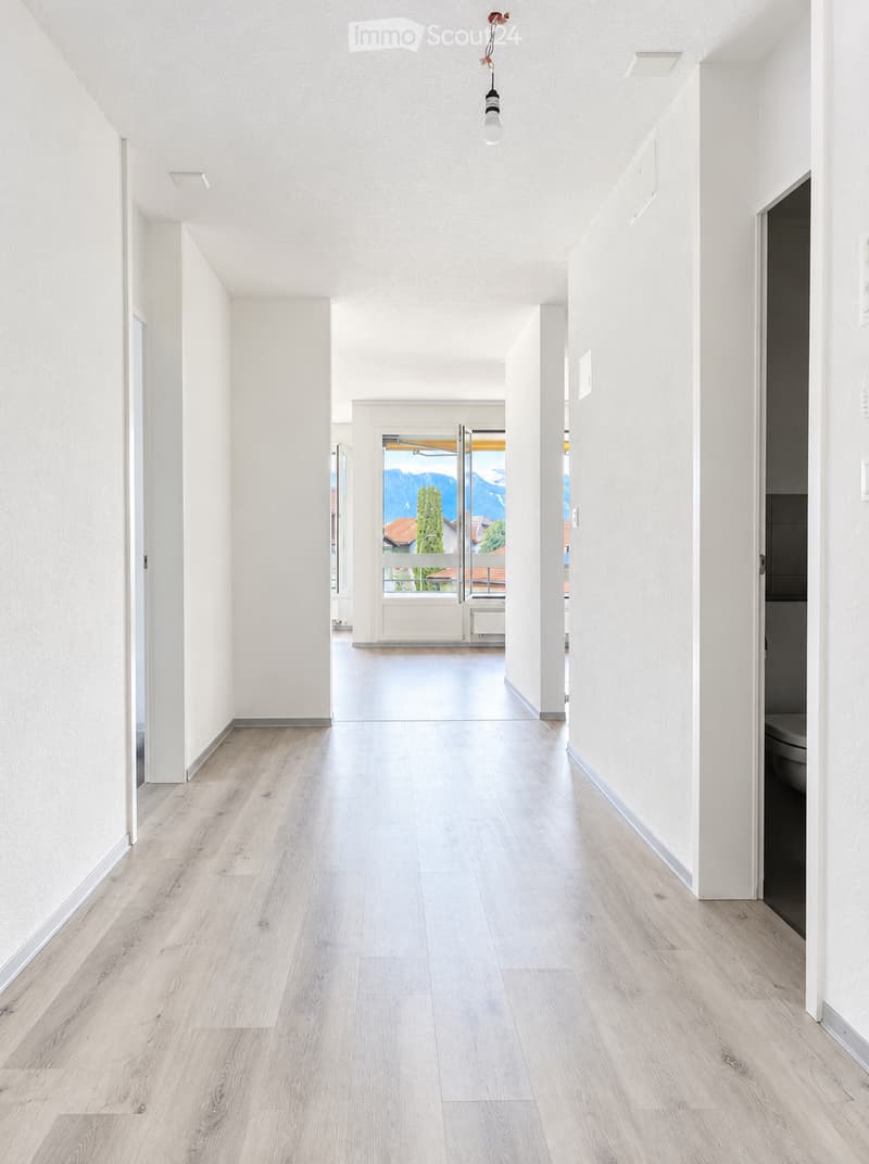 Bright, minimalist hallway with light wooden flooring and white walls, leading to an open doorway with a scenic mountain view.