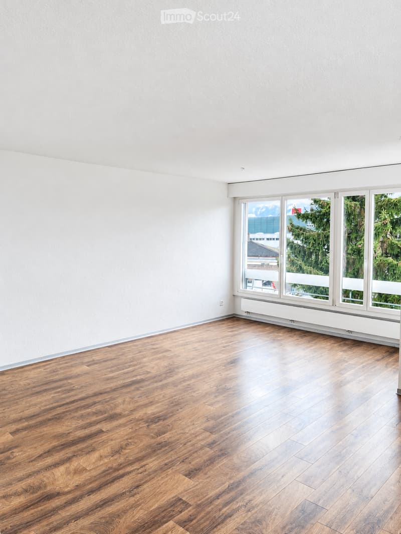 Empty room with light-colored walls and wooden flooring, featuring large windows with white frames and a radiator below them.