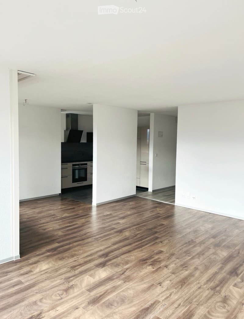 Empty room with wooden flooring and white walls, featuring an open kitchen area with cabinets and appliances visible in the background.