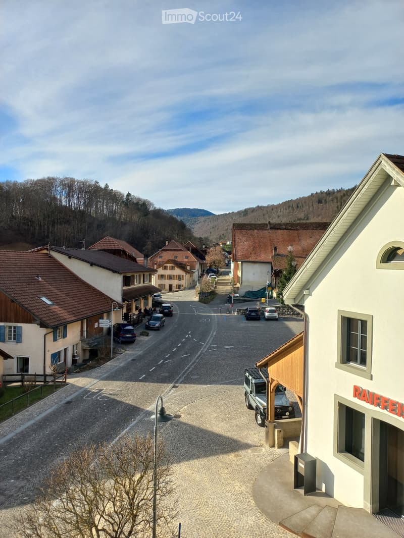 A small village street with traditional houses, parked cars, and a clear view of the surrounding hills and trees.