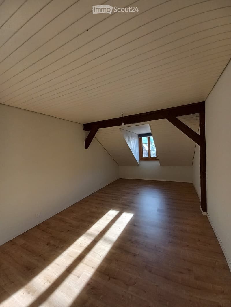 Empty attic space with wooden flooring, slanted ceilings, and exposed wooden beams. Sunlight streams in from a small window at the far end.