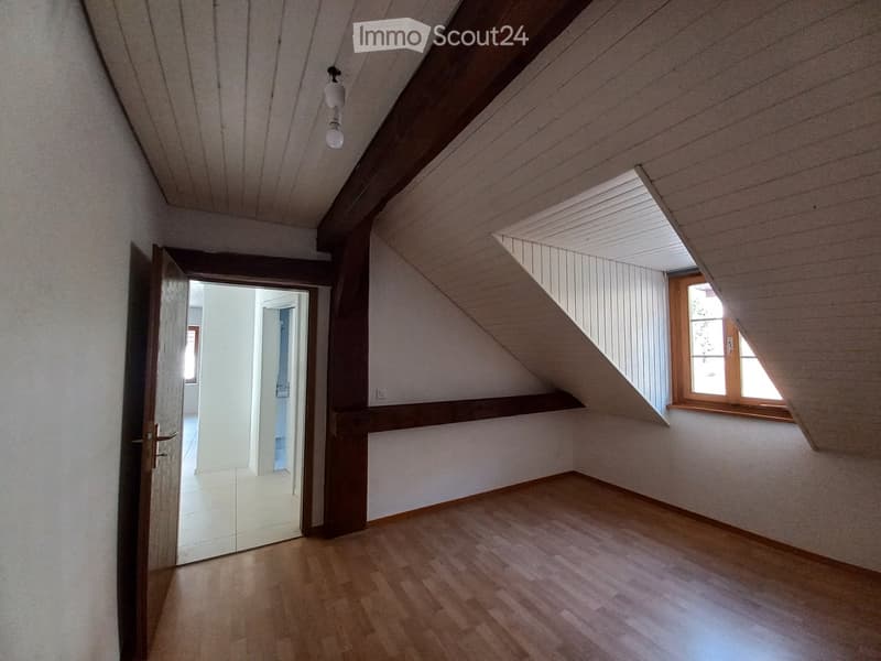 Empty attic room with wooden flooring, slanted wooden ceiling, and a single window letting in natural light. The room features a doorway leading to another space.