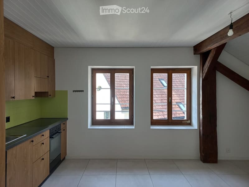Empty kitchen with wooden cabinets, green backsplash, tiled floor, and two windows overlooking a roof.
