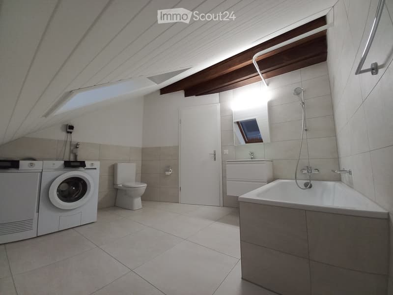 Modern bathroom with a bathtub, toilet, washing machine, sink, and mirror. Features skylight windows and a sloped ceiling.
