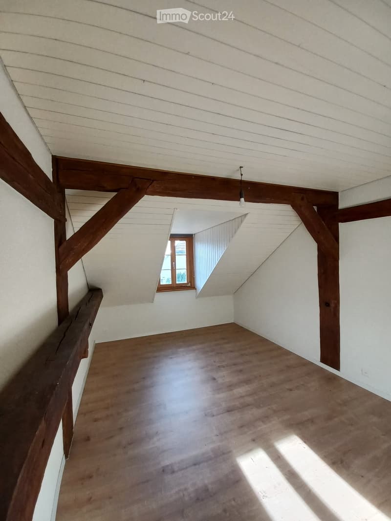 Empty attic space with wooden beams, slanted ceiling, and a small window letting in natural light.