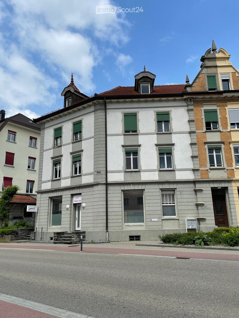 A multi-story building with a white facade, green shutters, and a red-tiled roof. The building has several windows and a central entrance with a sign reading "Aldora AG."