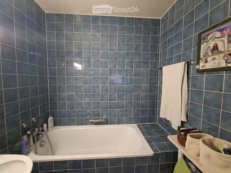 Bathroom with blue tiled walls and floor, a white bathtub, a towel hanging on a rack, a framed picture, and a small shelf with baskets and items.