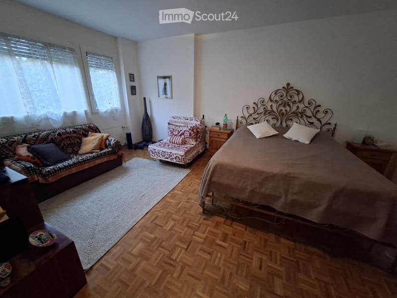 Cozy bedroom with wooden flooring, a large bed with an ornate headboard, a patterned sofa, a matching armchair, a nightstand, and a small table with a vase and picture frames on the wall.