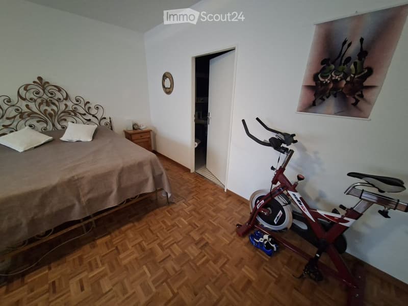 A furnished room with a bed, nightstand, exercise bike, and a picture frame on the wall.