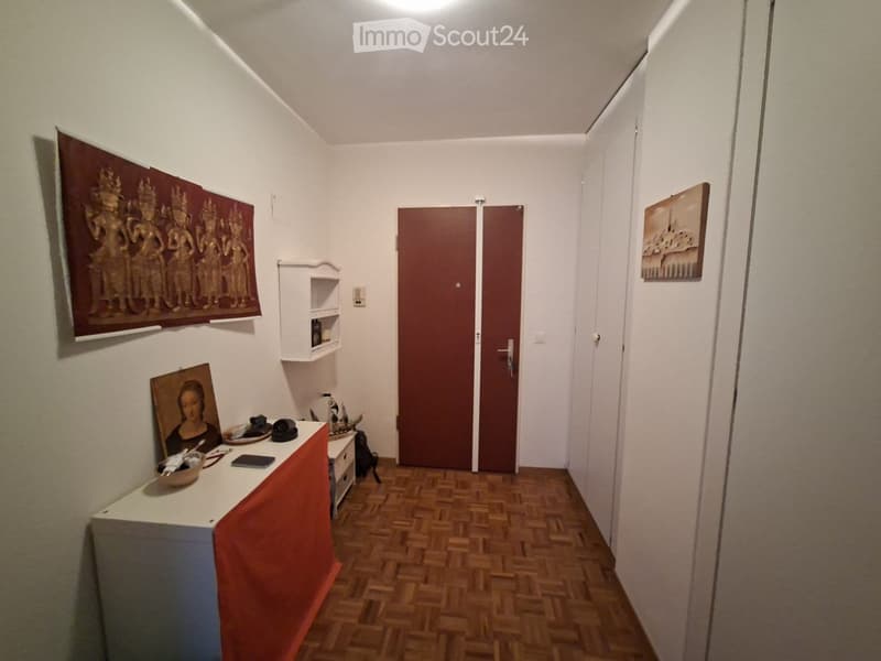 A narrow hallway with wooden flooring, white walls, and a double door at the end. There are small shelves and artwork on the walls, and a small table with various items on the left side.