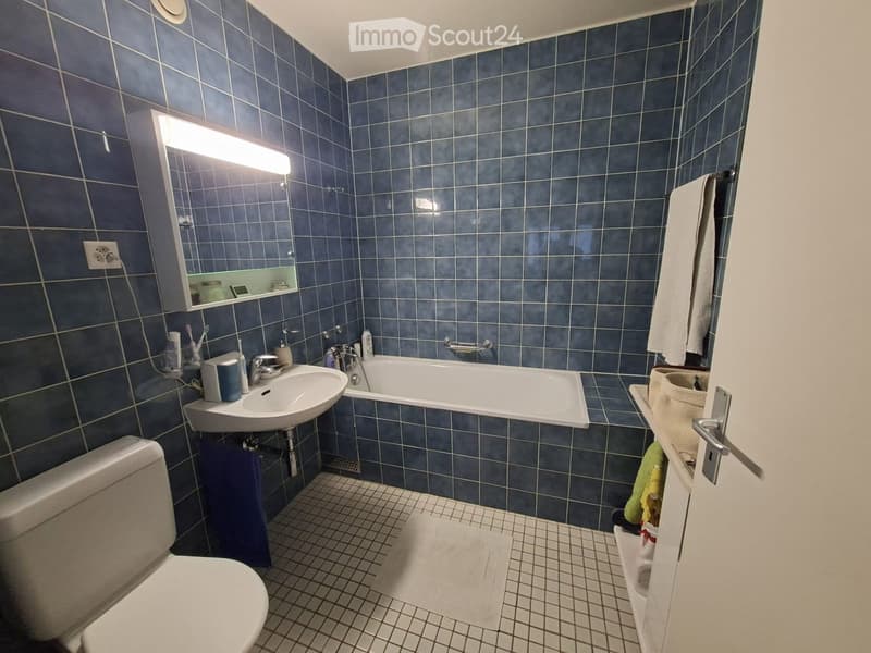 Bathroom with blue tiled walls and floor, featuring a toilet, sink, bathtub, mirror, and towel. The sink has a faucet and toothbrush holders, and a handrail is visible next to the bathtub.