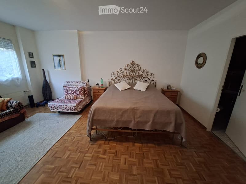 Cozy bedroom with wooden herringbone flooring, a large bed with ornate headboard, a patterned sofa, nightstands, and decorative items.