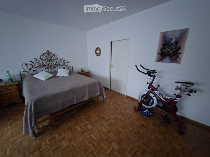 Bedroom with wooden flooring, a large bed with a decorative headboard, a nightstand, and an exercise bike in the corner.