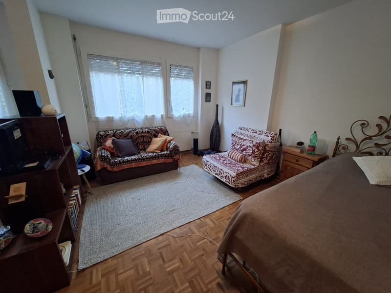 A furnished room with a bed, sofa, chair, small table, shelf, and a lamp. The room has wooden flooring and large windows with sheer curtains.