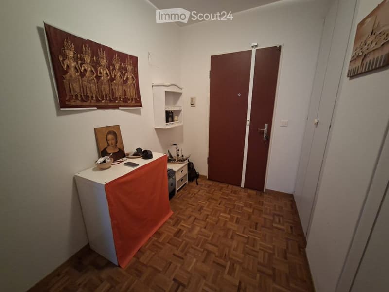 A narrow hallway with wooden flooring, a small table covered in an orange cloth, a shelf with decorative items, and framed artwork on the walls.