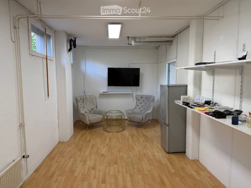 A small, modern room with light wooden flooring, white walls, and a mounted television. It features two armchairs, a small round table, a refrigerator, and a counter with office supplies.