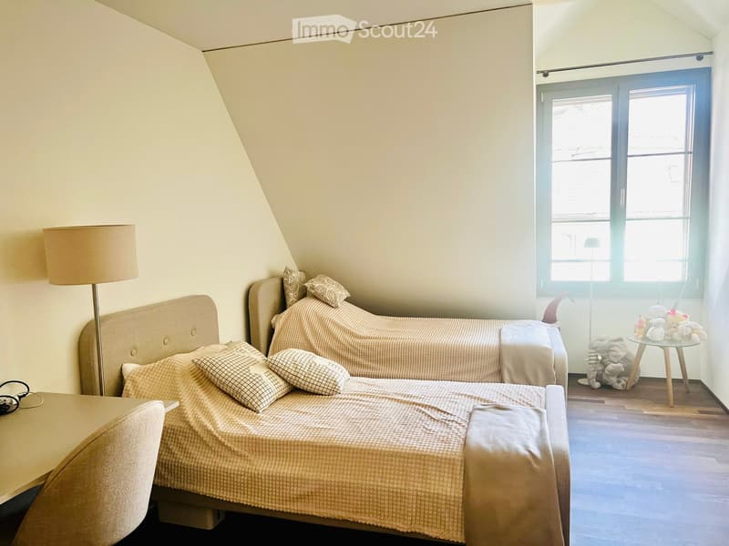 Cozy attic bedroom with two single beds, a small table, a floor lamp, and a few toys on a stool.