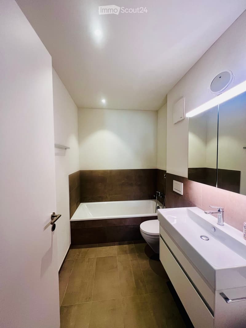Modern bathroom with a bathtub, toilet, sink, and mirror. Features a sleek design with light-colored tiles and a dark accent wall.