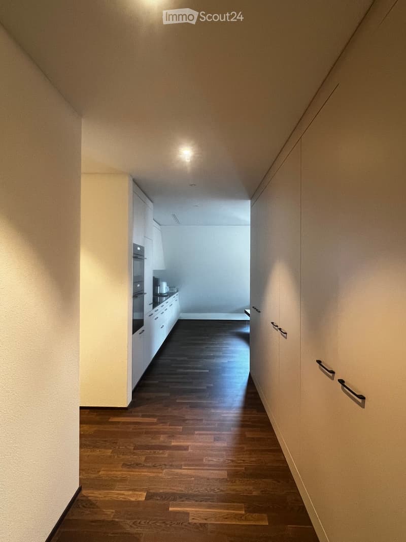A narrow hallway with wooden flooring and white walls, leading to a small kitchen area with white cabinets and a countertop.