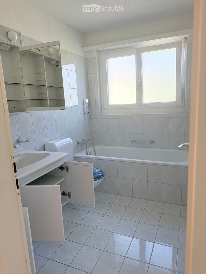 Modern bathroom with a white sink, toilet, bathtub, and mirrored cabinet. The room features tiled walls and floor, with a window providing natural light.