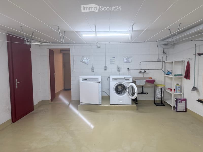Utility room with white brick walls, concrete floor, washing machine, dishwasher, sink, shelf with cleaning supplies, and a towel hanging on the wall.