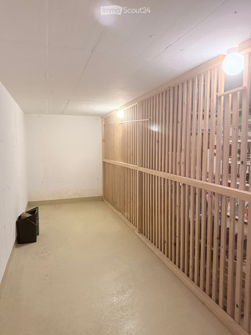 A narrow hallway with a wooden railing on one side, white walls, and a light-colored floor. A green trash bin is visible on the left side.