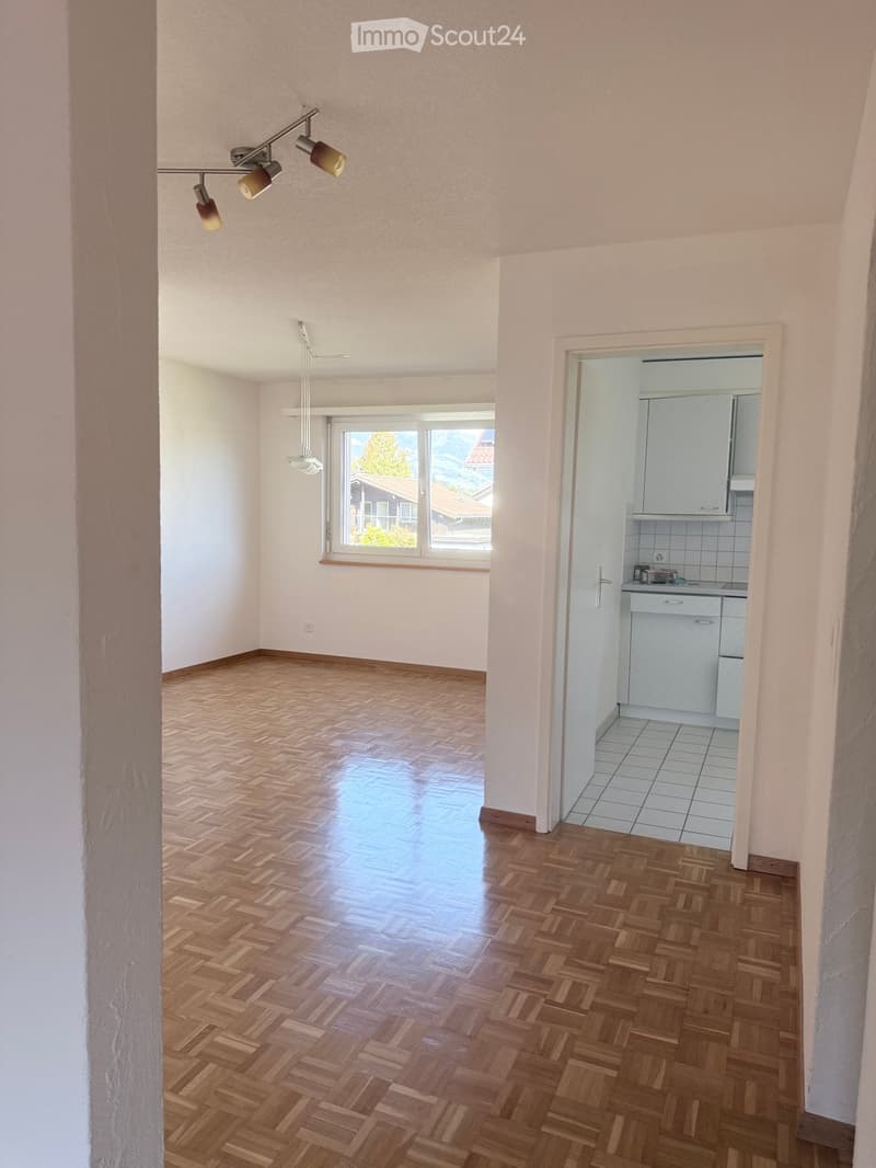 Empty room with wooden herringbone flooring, white walls, and a large window with a scenic view. Adjacent kitchen area visible with white cabinets and tiled flooring.