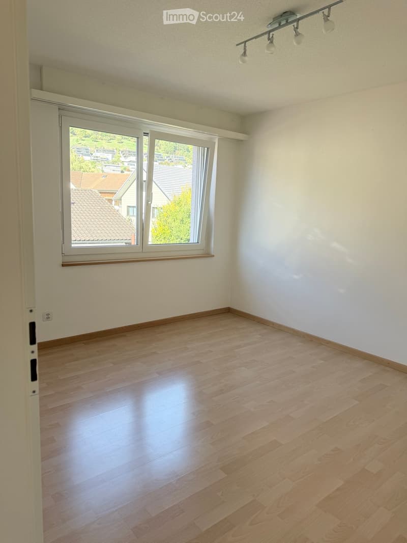 Empty room with light wooden flooring, white walls, and large windows offering a view of the neighborhood.