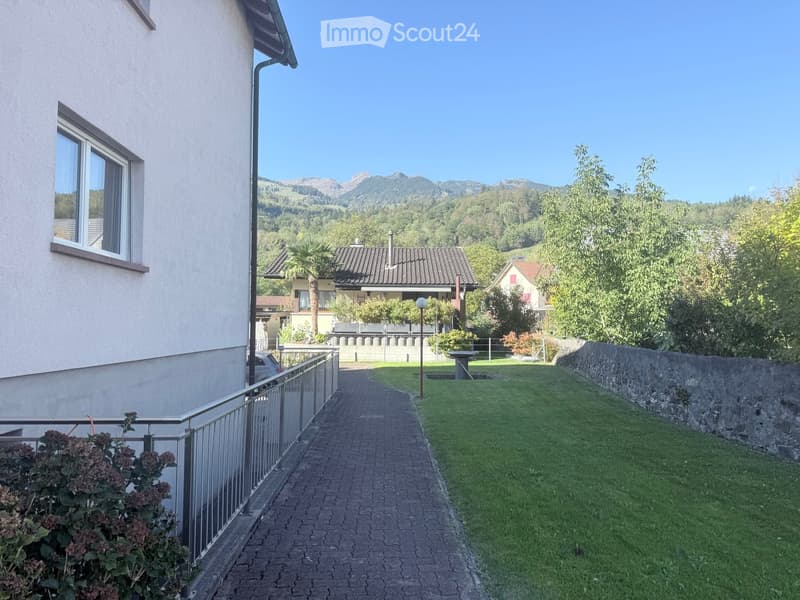 A well-maintained garden area with a paved pathway, metal railing, and a view of surrounding greenery and mountains.