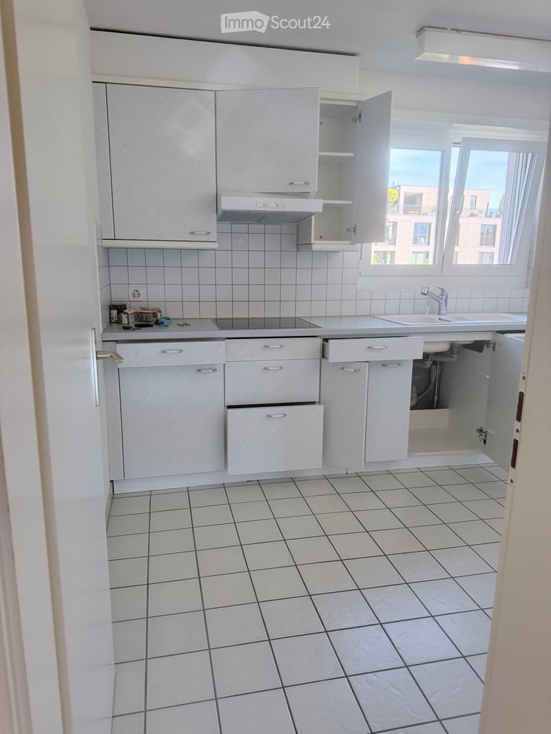Modern kitchen with white tiled walls and floor, white cabinets, sink, and faucet. The cabinets have silver handles and the sink is positioned under a window with a view of the outside.