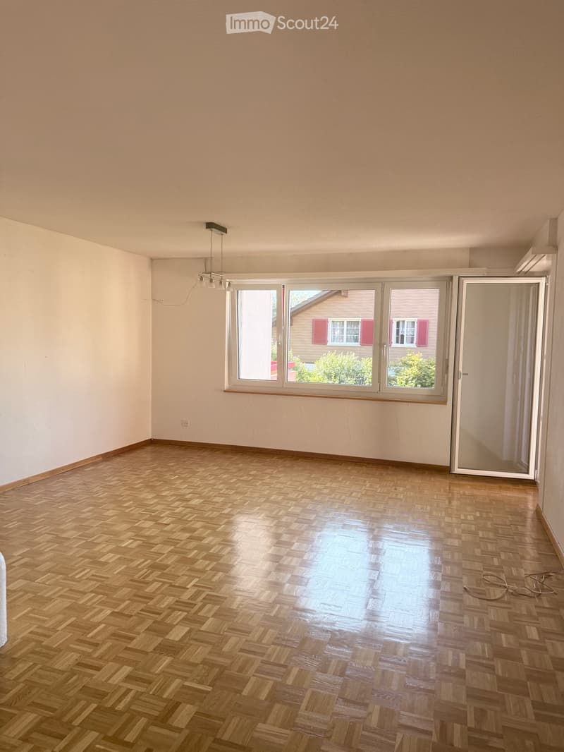 Empty room with wooden herringbone flooring, white walls, large windows, and a ceiling light fixture.