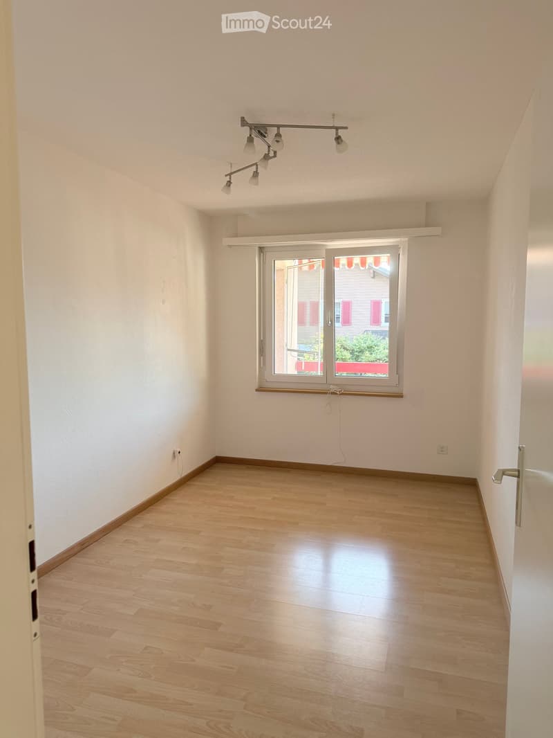 Empty room with light wooden flooring, white walls, and a large window with blinds. The room features a modern ceiling light fixture and a door on the right side.