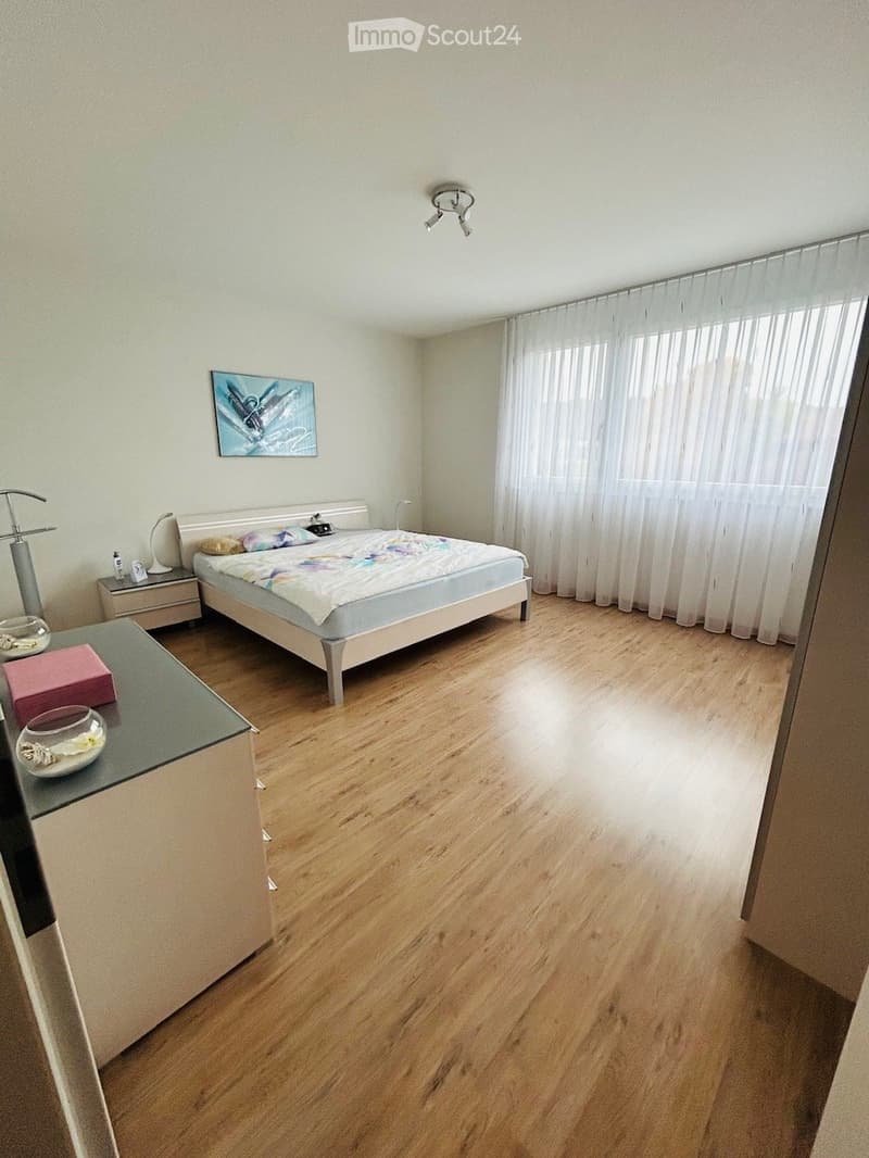 Bright bedroom with wooden flooring, a bed with colorful bedding, a nightstand with a lamp, a small table with decorative items, and a large window with white curtains.