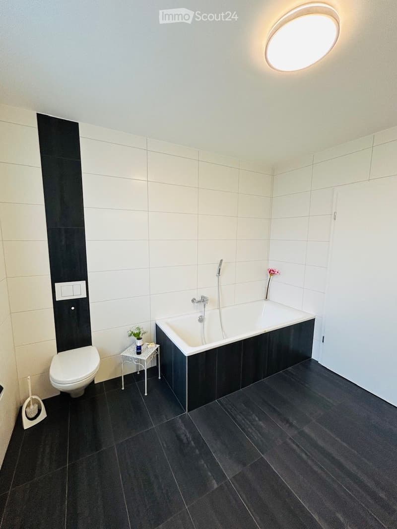 Modern bathroom with white tiles, black accents, bathtub, toilet, small table with flower vase, and a round ceiling light.