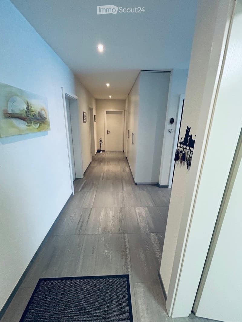 Clean, well-lit hallway with tiled flooring, white walls, and several doors leading to rooms. A small rug is placed near the entrance, and artwork and a key holder are visible on the walls.