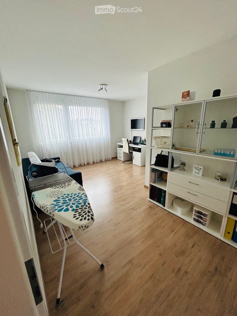 A well-lit room with wooden flooring, a sofa, an ironing board, a mirror cabinet, a desk with a laptop, and a chair.