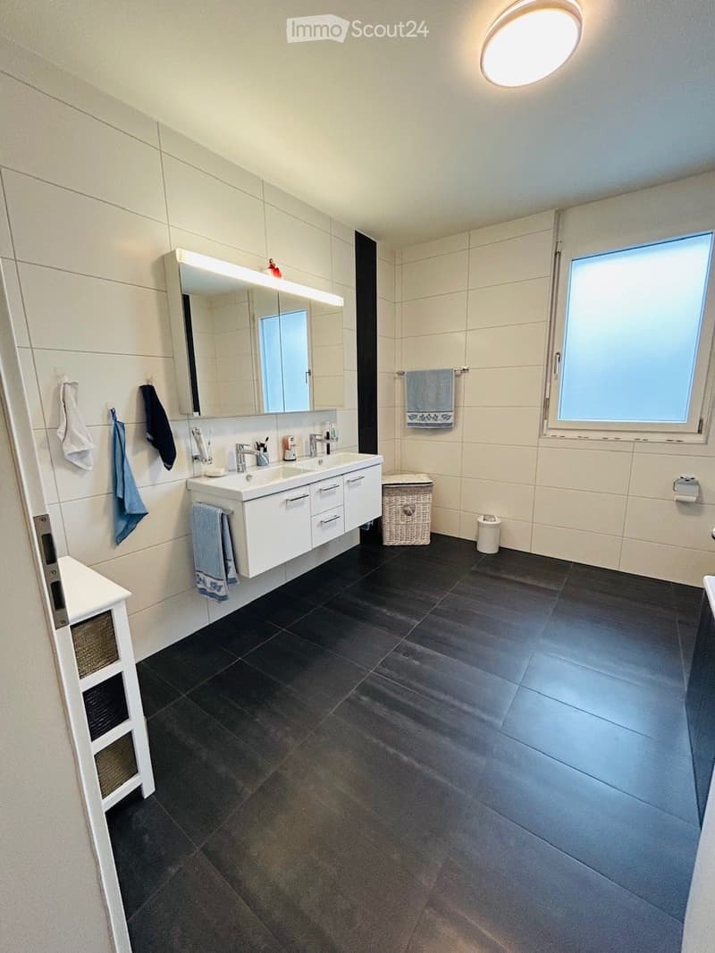 Modern bathroom with dark tiled floor, white tiled walls, double sink vanity, large mirror with built-in lighting, and various towels and accessories.