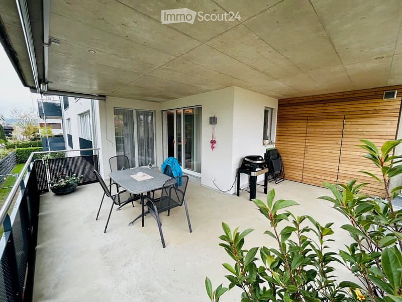 Modern balcony with a dining table, chairs, barbecue, and potted plants, featuring large glass doors and wooden paneling.