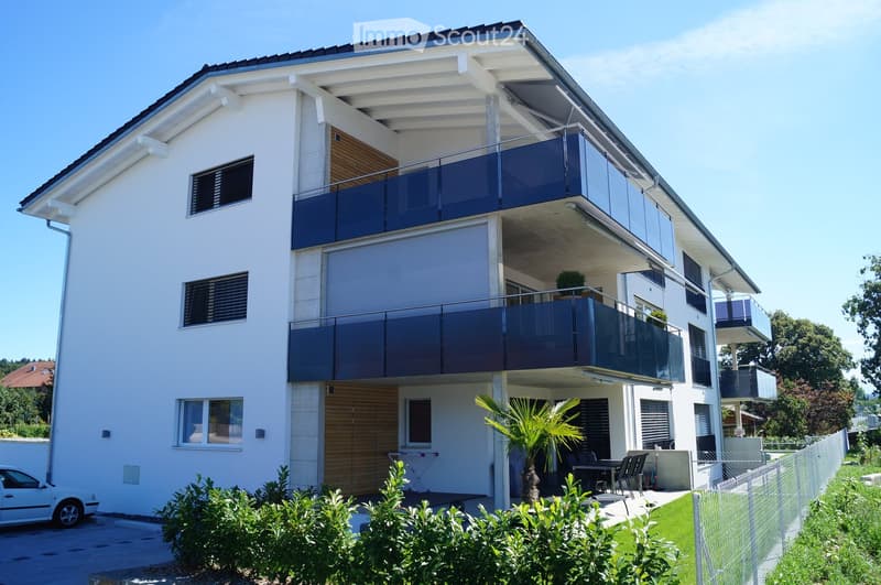 Modern two-story house with white exterior, large windows, balconies, and a small garden area with outdoor furniture.