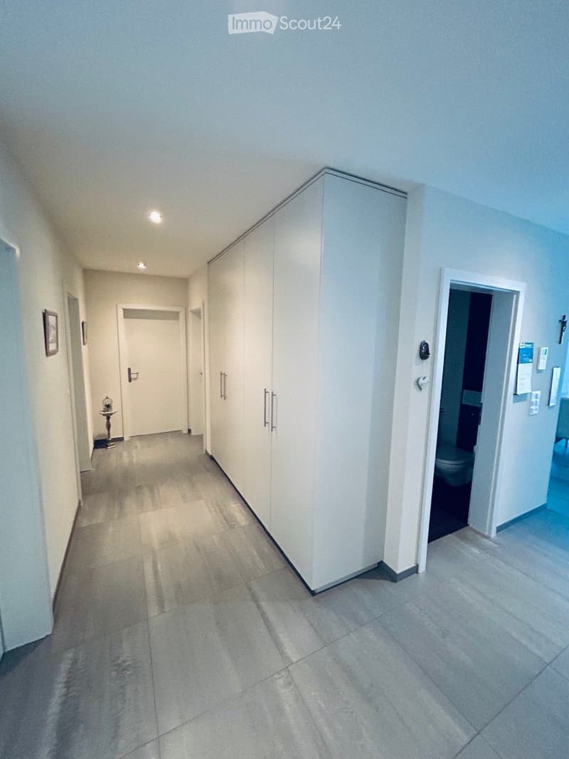 Clean, modern hallway with tiled flooring, white walls, built-in storage units, and recessed lighting. Doors lead to other rooms, including a visible bathroom.