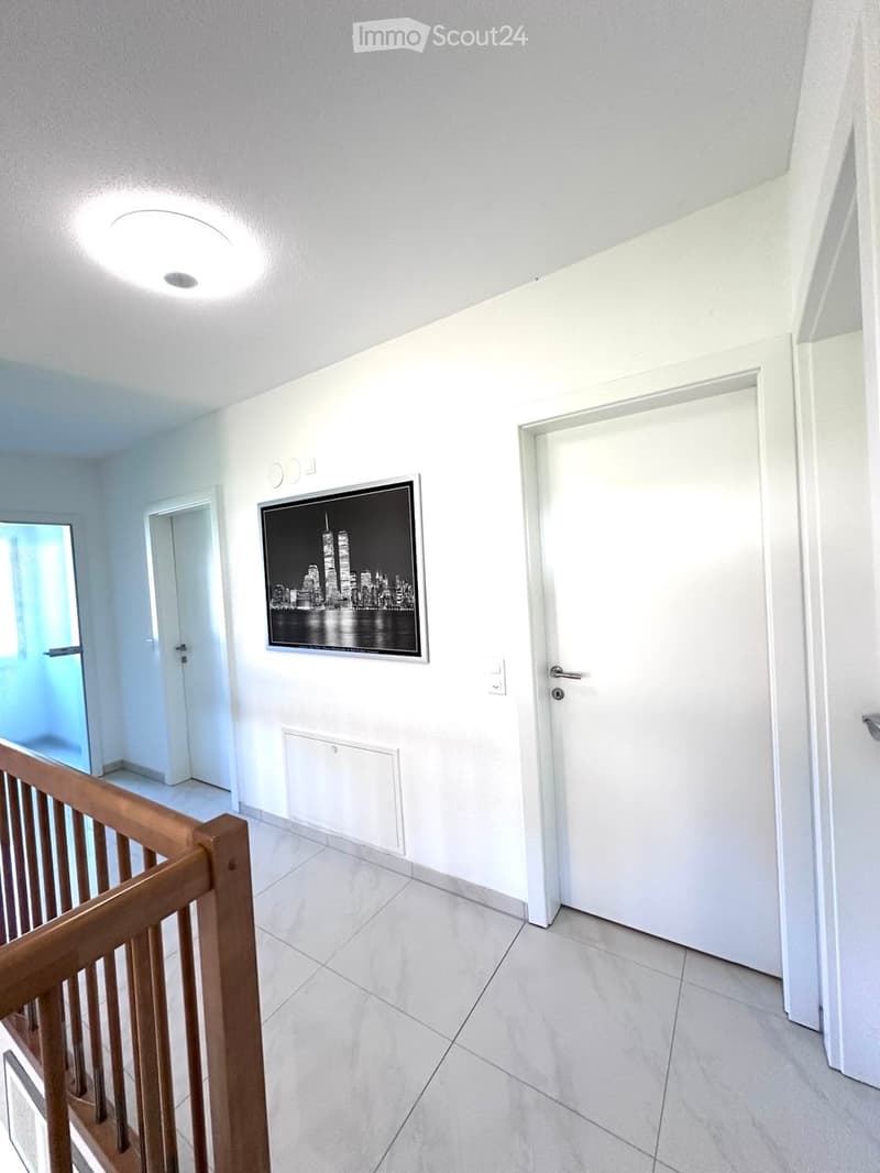 A modern, minimalistic hallway with white walls, tiled flooring, and a wooden railing leading to an upper level. A large framed cityscape artwork is mounted on the wall, and several doors are visible.