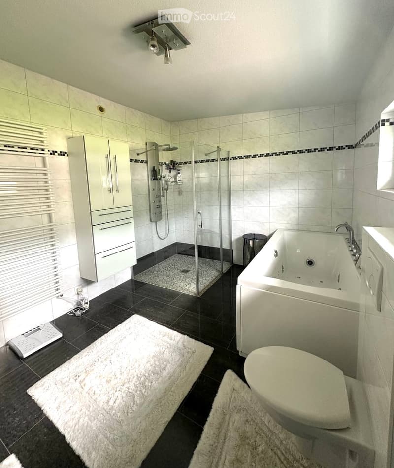 Modern bathroom with a bathtub, toilet, glass-enclosed shower, white cabinet, and a large mirror. The floor is tiled in dark gray, and there are two white rugs placed in front of the bathtub and toilet.