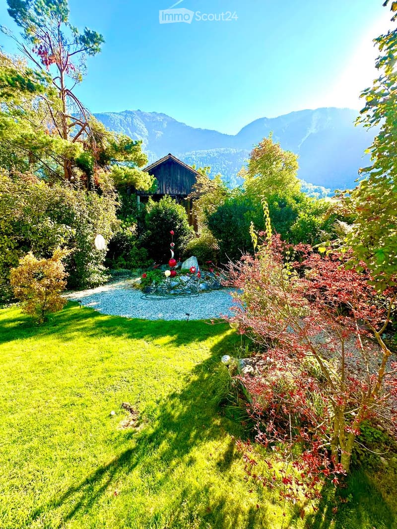 A well-maintained garden with a gravel pathway, a small pond with decorative elements, lush green grass, and various trees and shrubs, with a wooden house in the background and mountains visible in the distance.