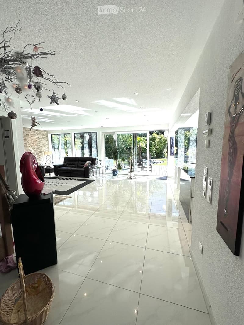 Modern, spacious living room with white tiled floors, large windows, and a black leather sofa. A red abstract sculpture sits on a black stand, and a wicker basket is placed near the entrance.
