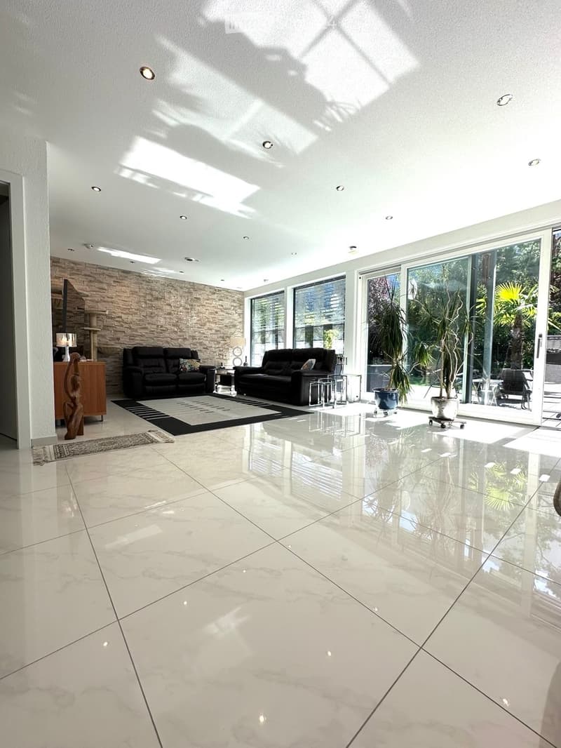 Modern living room with polished marble floors, large glass windows, two black leather sofas, a coffee table, side tables, and a potted plant near the window.