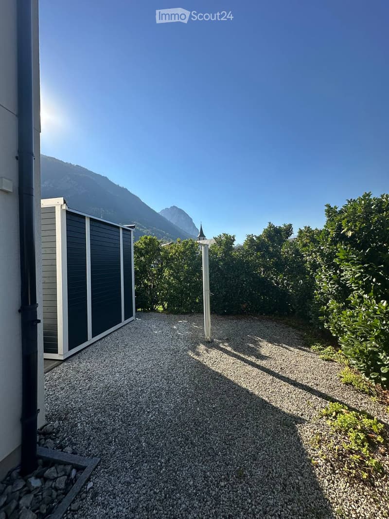Gravel pathway leading to a white shed, surrounded by greenery and mountains in the background.