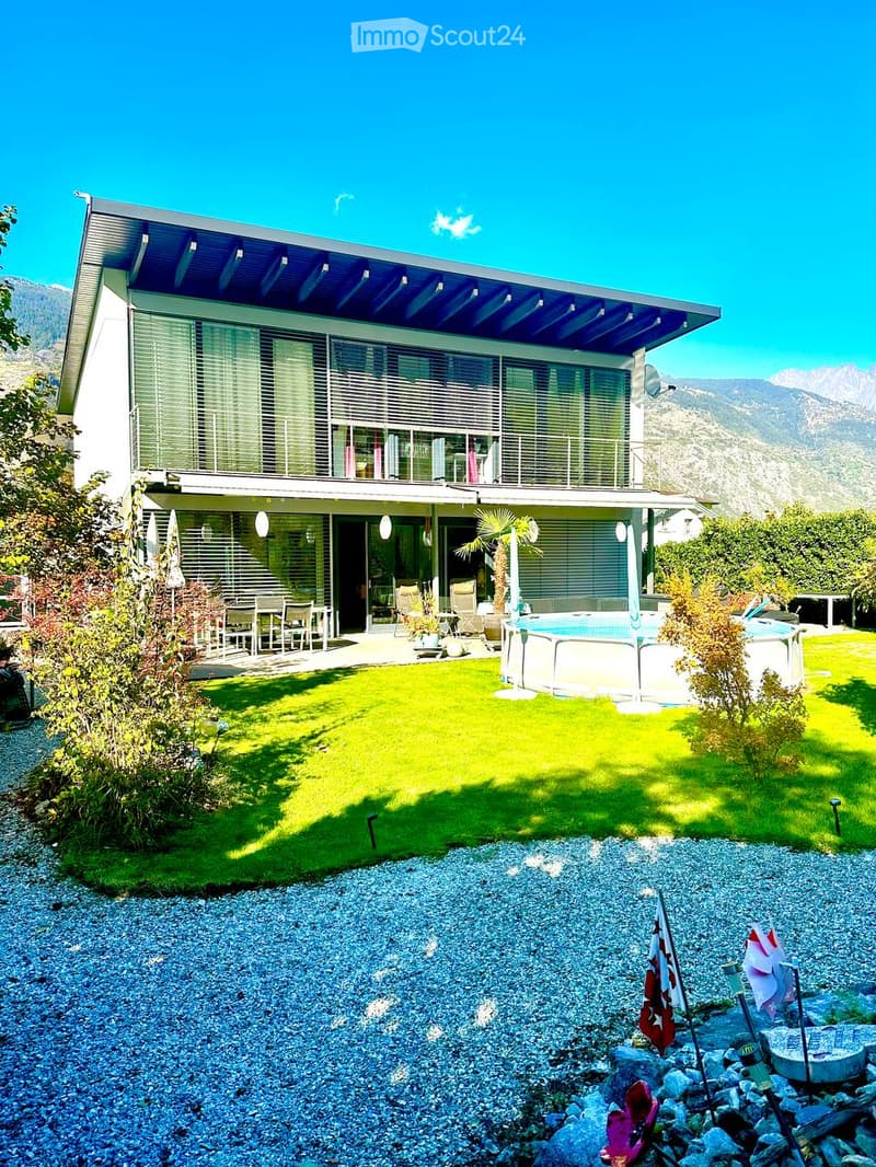 Modern two-story house with large windows, a balcony, and a swimming pool in the garden, surrounded by greenery and a mountain view.