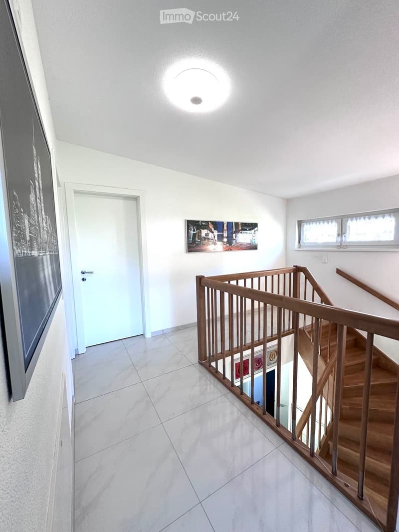 Modern hallway with white tiled flooring, wooden staircase with glass railing, white walls, and a round ceiling light. A framed picture hangs on the wall, and a window provides natural light.