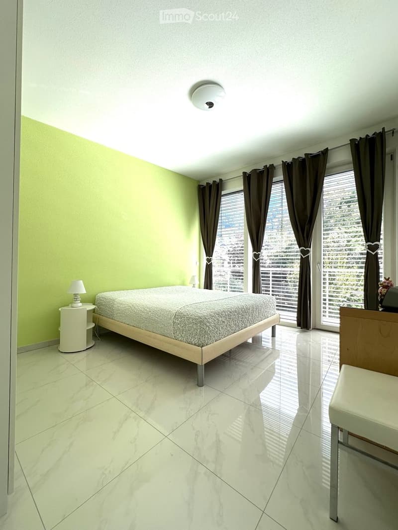 Modern bedroom with a large bed, nightstands, lamps, and a view through large windows with blinds and curtains.
